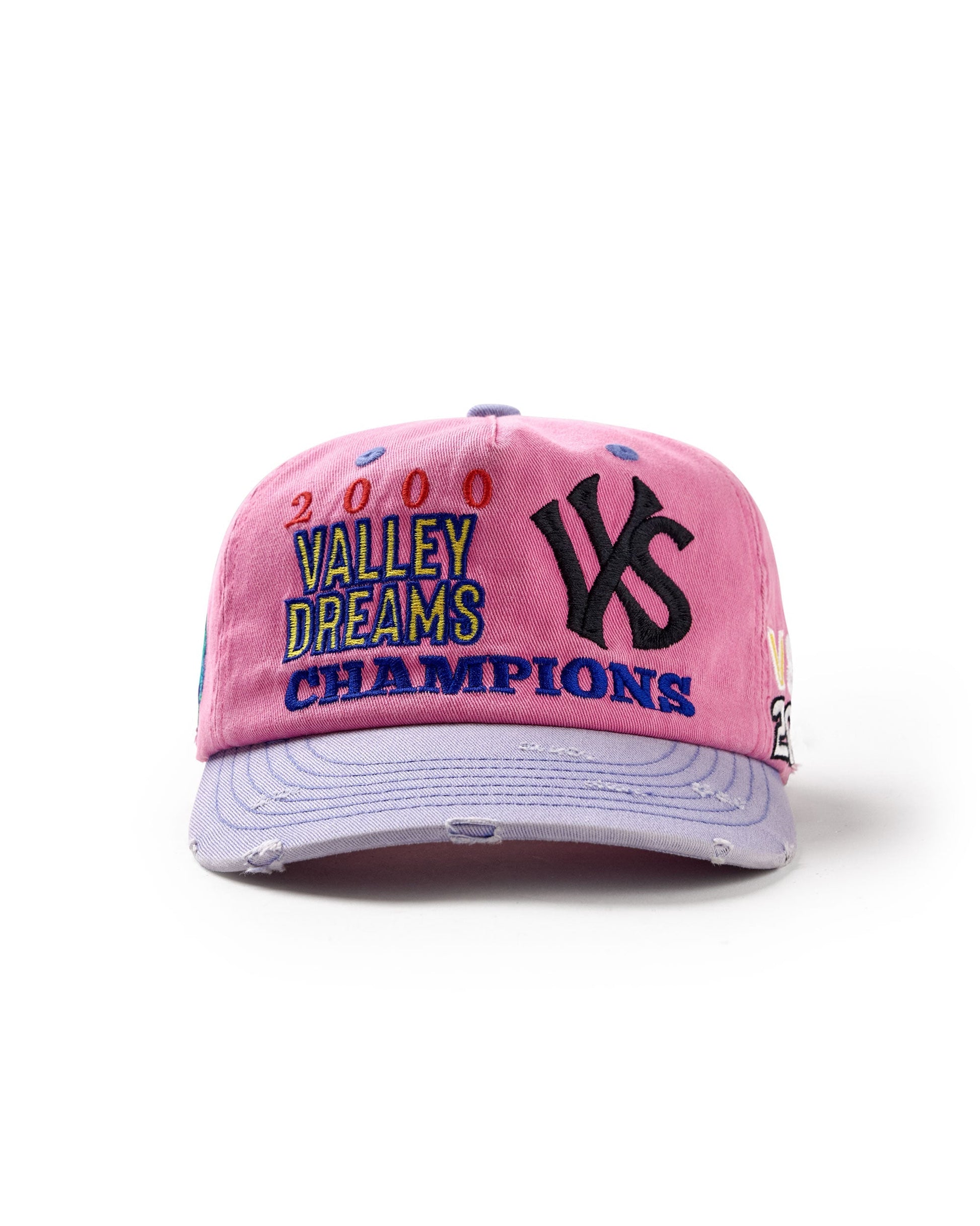 A pink distressed baseball cap featuring embroidered text that reads '2000 VALLEY DREAMS CHAMPIONS' in multicolor lettering, alongside a large black embroidered 'VS' logo on the front. The cap has a light lavender brim with distressed details and is photographed on a clean white background.