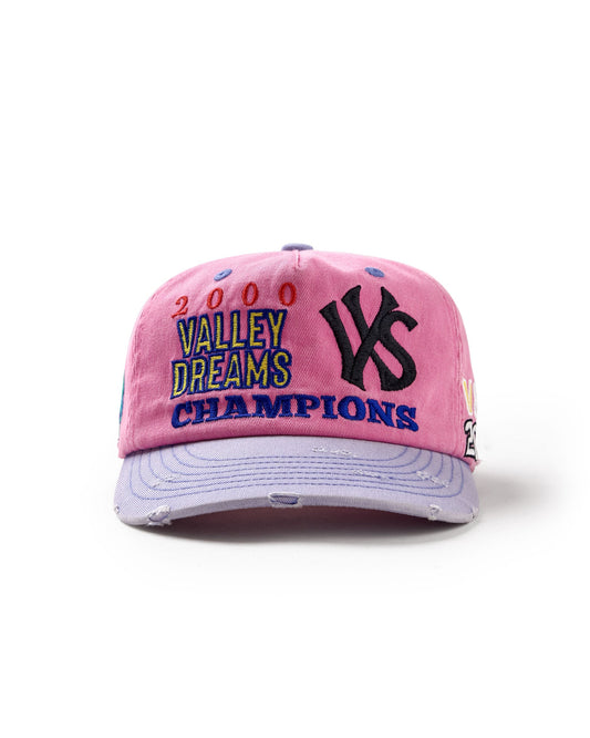 A pink distressed baseball cap featuring embroidered text that reads '2000 VALLEY DREAMS CHAMPIONS' in multicolor lettering, alongside a large black embroidered 'VS' logo on the front. The cap has a light lavender brim with distressed details and is photographed on a clean white background.